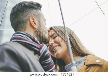 Beautiful young couple standing close to each other under an umbrella on a rainy day - Man kissing his girlfriend on the forehead - Boyfriend and girlfriend having tender moments in the rain