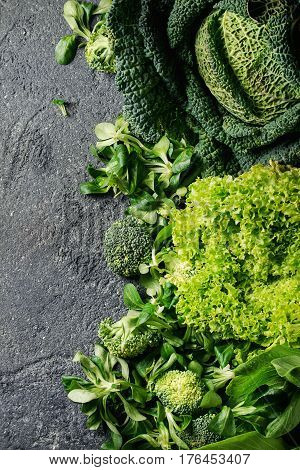 Variety of raw green vegetables salads, lettuce, bok choy, corn, broccoli, savoy cabbage as frame over black stone texture background. Space for text