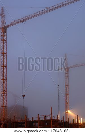 Two cranes on the construction site, unfinished house, foggy evening twilight, building lighting.