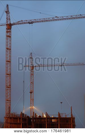 Two cranes on the construction site, unfinished house, foggy evening twilight, building lighting.