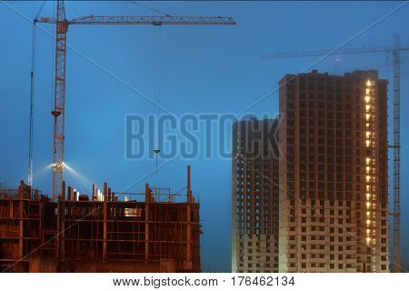 Cranes on a large construction site, unfinished multi-storey houses, foggy evening twilight.