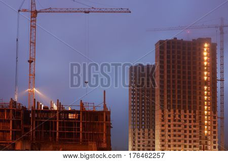 Construction crane on the site, unfinished multi-storey houses, fog covers the upper floors, evening twilight.