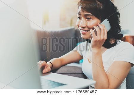 Portrait of young smiling Asian woman working at modern laptop home, wearing white tshirt and talking on smartphone.Horizontal blurred, flare