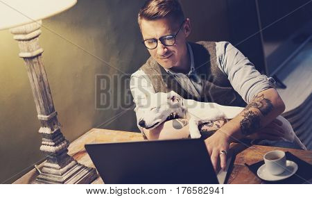 Handsome tattooed man in eyeglasses working at home on laptop while sitting at the wooden table with cute dog sleeping on his hands.Adult guy using modern computer for surfing web