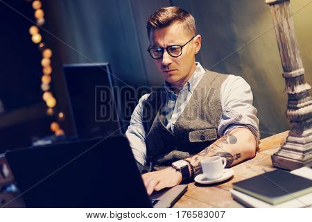 Pensive tattooed man in eyeglasses working at home on laptop while sitting at the wooden table.Using modern computer for research new startup ideas.Concept of business people work.Blurred background