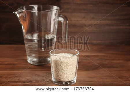 Glass of rice and measuring jug with water on wooden table