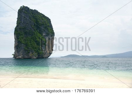 Big high rock cliff filled with green vegetation surrounded by turquoise blue colored ocean water next to a tropical white sand beach with horizon view at midday Krabi Thailand.