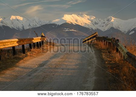 Old road with rusty road fences pointing towards snowy mountains in background in warm orange light of sunset