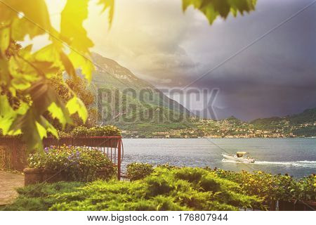 Beautiful toned waterscape of coastline of italian Varenna city with focus on mountain on background leaf on foreground trees and dramatic cloudy sky before the rain
