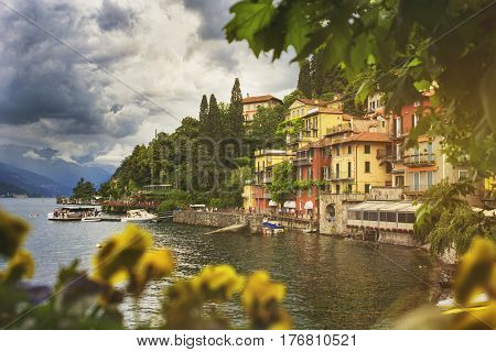 Beautiful cityscape with a Como lake coastline of italian Varenna city with colorful building big trees and dramatic sky before the rain with a bokeh flowers on foreground