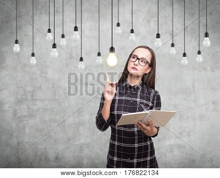 Portrait of a nerdy girl with a book standing near a conccrete wall with multiple light bulbs hanging on their wires.