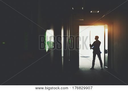 Silhouette of boss or businessman in formal suit standing in dark office corridor interior with reflections and green lights and holding katana sword like japanese samurai warrior ready to fight