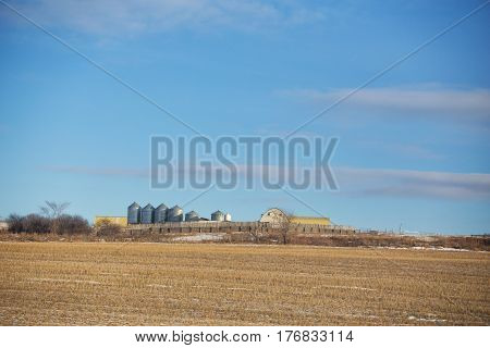 Steel grain storage bins and roof tops of buidings visible behind a long rustic wooden fence by a harvested field in a rural countryside landscape
