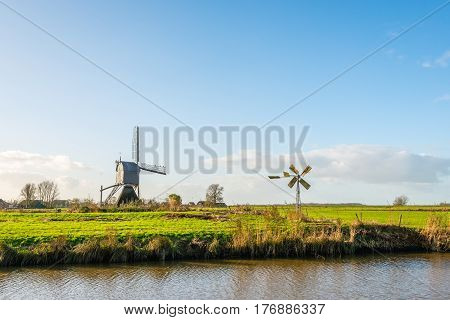 Back of a wooden hollow post mill and a small metal polder mill next to each other in a Dutch polder landscape on a sunny day in autumn.
