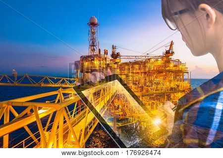 Double exposure of businesswoman in hipster shirt and wearing glasses working with tablet and oil and gas refinery industry plant in the night as Energy and Technology concept.