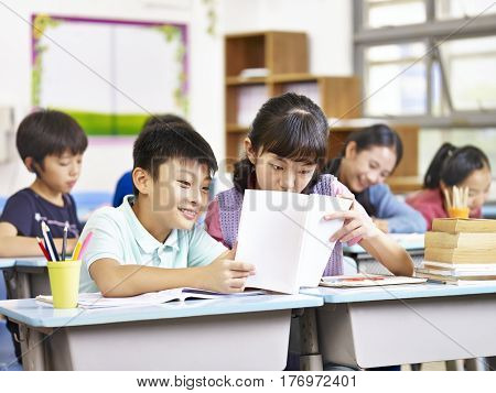 asian elementary schoolgirl and schoolboy sharing a book in classroom.