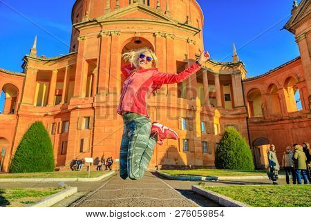 Happy Woman Jumping At Madonna Of San Luca Sanctuary. Popular Landmarks In Bologna, Italy. Blonde Fe