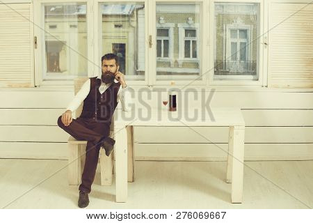 Man Sitting On Wooden Chair At Table With Bottle And Glass Of Red Wine. Brutal Hipster With Beard An