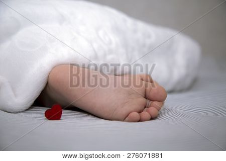 The Baby Sleeps Under A Blanket, Feet Out With A Small Red Heart For The Holiday