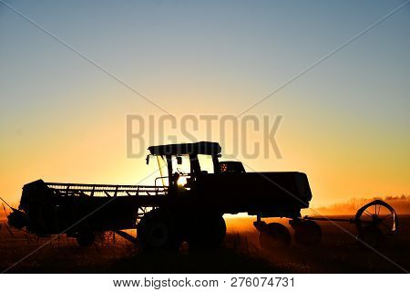 A Silhouette Image Of Agricultural Equipment In A Field At Sunset.