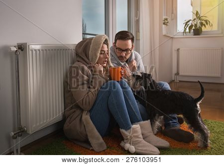 Young Couple In Jacket And Covered With Blanket Sitting On Floor Beside Radiator With Dog And Trying