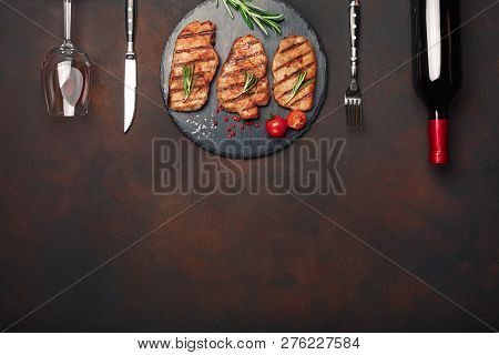 Grilled Pork Steaks On Stone With Bottle Of Wine, Wine Glass, Knife And Fork On Rusty Background