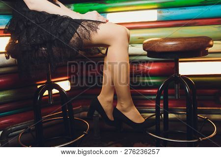 A Close Up Image Of The Legs Of A Young Woman In A Dress Sitting Ona Barchair In Black Heels.