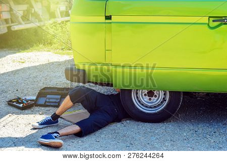 Young Male Hipster Lying Beneath His Old Timer Camper Van Trying To Repair It Beside Road