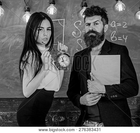 Discipline Concept. Man With Beard Hold Book And Sexy Girl Teacher Holds Alarm Clock, Chalkboard On 