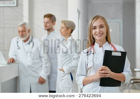 Young Female Doctor Standing With Tablet And Looking At Camera. Her Colleagues Standing Behind Her A