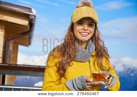 Woman drinking warm tea in the rustick wooden terrace on mountain, alpine view, snow on hills. 