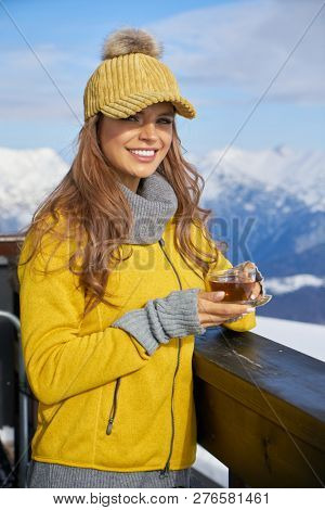 Woman drinking warm tea in the rustick wooden terrace on mountain, alpine view, snow on hills. 