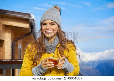 Woman drinking warm tea in the rustick wooden terrace on mountain, alpine view, snow on hills. 