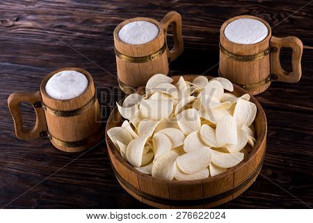 Three Oak Mugs With Beer Inside. They Stand Near An Oak Tray In Which Are Potato Chips. On Dark Wood