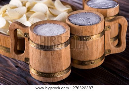 Three Oak Mugs With Beer Inside. They Stand Near An Oak Tray In Which Are Potato Chips. On Dark Wood