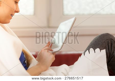 Young Woman Lying On Sofa Under Warm Blanket, Relaxing Using Tablet, Reading News And Checking Socia