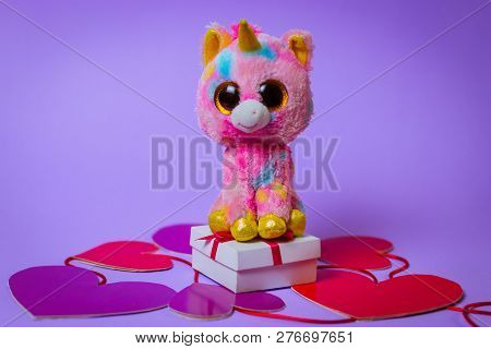Toy Pink Unicorn Sits On A White Gift Box With A Red Ribbon On Paper Hearts On A Purple Background