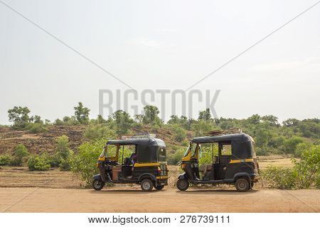 Two Black And Yellow Indian Motor Rickshaws Stand On An Asphalt Road Against The Background Of A Hil