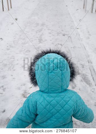 Back Of Small Child In Blue Turquoise Color Down Jacket Walking Out From The Camera Look Ahead At St
