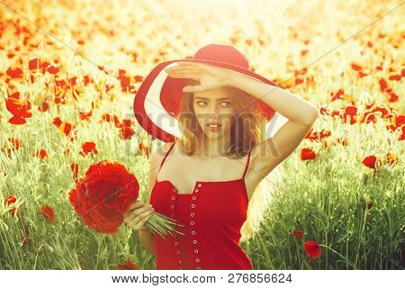 Girl With Long Curly Hair In Red Dress And Retro Hat Hold Flower Bouquet In Field Of Poppy Seed On G