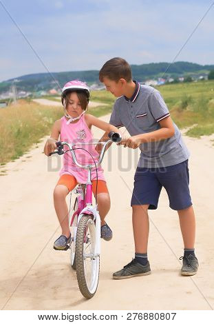 Youth Brother Teaching His Younger Sister To Ride A Bike. Little Girl In A Pink Protective Helmet On