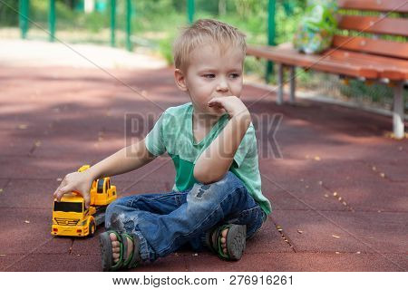 Little Boy With Blue Eyes Sits On The Cover Of Children Playground With A Toy - Yellow Excavator. Bl