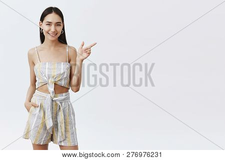 Studio Shot Of Charming Asian Dark-haired Female In Trendy Matched Top And Shrits, Pointing Right Wi