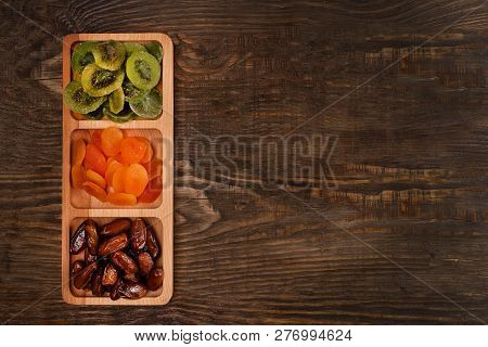 Dates, Dried Apricots And Kiwis In A Compartmental Dish On A Dark Wooden Table.