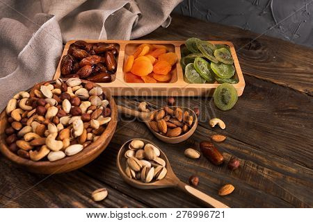 Dates, Dried Apricots And Kiwis In A Compartmental Dish And Assortment Of Nuts In Wooden Bowl On A D