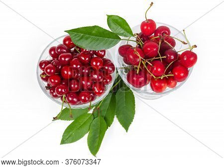 Fresh Sour Cherries And Sweet Cherries In The Two Glass Cups, Leaves Of Cherry Tree On A White Backg