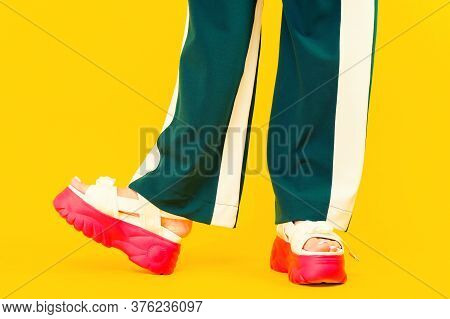 Women's Legs In Sport Sandals With Red Soles And Green Pants With Stripes On A Yellow Background.