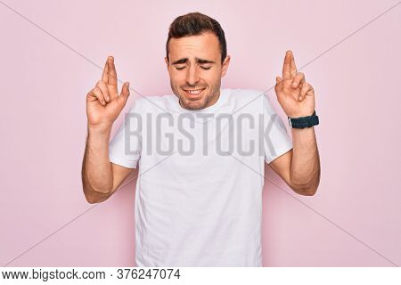 Handsome man with blue eyes wearing casual white t-shirt standing over pink background gesturing finger crossed smiling with hope and eyes closed. Luck and superstitious concept.