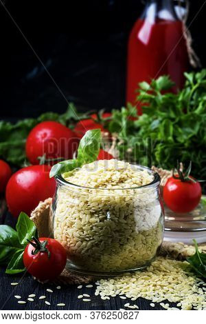 Italian Uncooked Pasta Riso Or Orzo In A Glass Jar, Tomatoes And Green Basil, Black Background, Sele