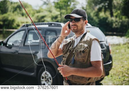 Young Fisherman Fishing On Lake Or River. Busy Guy Holding Fishing Rod And Talking On Smartphone. St
